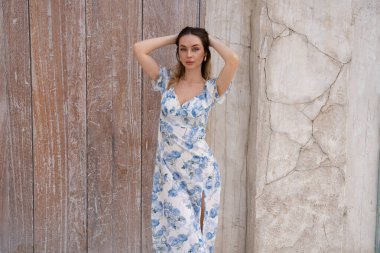 Açık mavi çiçekli güzel bir kadın ahşap kapının yanında poz veriyor, yumuşak güneş ışığı ve nötr renkler huzurlu ve zarif bir his yaratıyor.. 