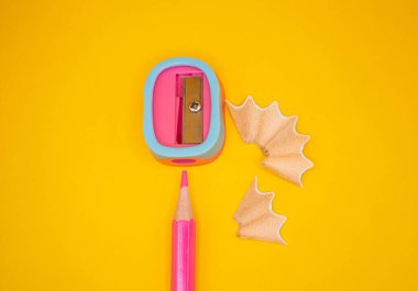 Renkli bileyicili pembe kalem ve parlak sarı arka planda kıvrımlı talaşlar. Eğitim, yaratıcılık ve okul temalı görseller için ideal. 