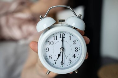 Bir el, sabah 6: 00 'yı gösteren, bulanık yatak odası arka planına sahip, sabah rutinini ve uyanma saatini simgeleyen beyaz analog bir çalar saati tutuyor..