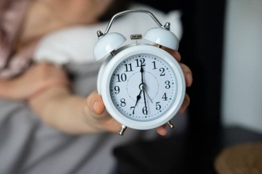 Sabahın erken saatlerinde elinde klasik beyaz alarmlı bir saat tutan, uyuyan bir bireyin bulanık arka planına sahip, uyanma hissini ve sabah rutinini aktaran bir kişi..