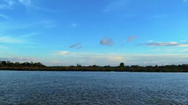 Pofuduk bulutlarla dolu canlı mavi bir gökyüzünün altında akan huzurlu nehir doğa severler ve seyahat meraklıları için sakin ve rahatlatıcı bir atmosfer yaratıyor.