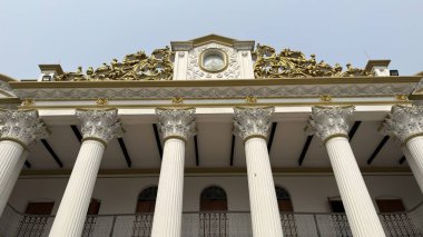 Karmaşık altın süslemeleri, sağlam beyaz sütunlar ve temiz gökyüzüne karşı merkezi bir saat içeren görkemli bir neoklasik bina..