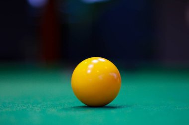 Yeşil bir masada duran parlak sarı bir bilardo topunun yakın çekimi, sığ bir alan derinliğinde yakalanmış. Parlak yüzey, yumuşak yansımalar ve bulanık arka plan spor atmosferi..
