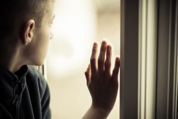 Печальный мальчик смотрит со стороны, держа стеклянное окно — стоковое фото