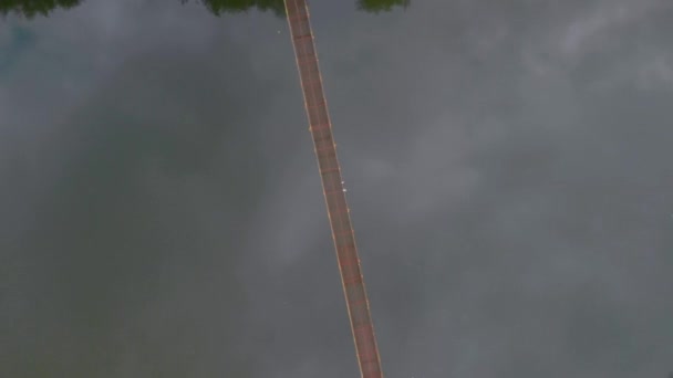 Дрон обертається над підвісним мостом з двома білими птахами на ньому над невеликою річкою в літній хмарний день з легким дощем — стокове відео