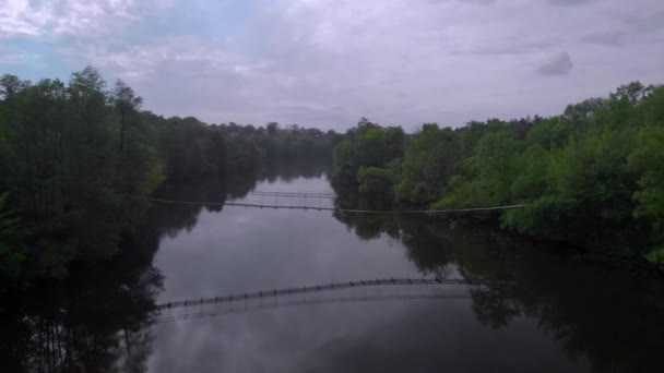 Yazın bulutlu ve hafif yağmurlu bir günde, küçük bir nehrin üzerindeki asma köprünün hava görüntüsü. — Stok video