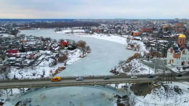 Беспилотник пролетает над автомобилями, проезжая небольшой мост через замерзшую маленькую реку и церковь в европейском городе — стоковое видео