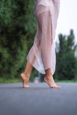 Akan bir elbiseyle asfalt yolda yürüyen çıplak ayaklı kadın. Özgürlük, yolculuk, farkındalık, denge ve yavaş yaşam kavramı. Açık hava minimalist sahnesi ve sığ alan derinliği.