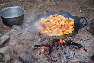 Açık bir kamp ateşinde metal bir tavada pişen kızarmış patates dilimleri. Kamp, hayatta kalma aşçılığı ve geleneksel kırsal yaşam tarzını temsil eden ateş, duman ve kırsal yemek takımı ile açık hava gıda hazırlığı.