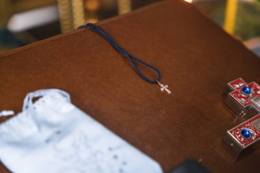 Ahşap bir yüzeye uzanan mavi ipli küçük metal bir haç kolyesinin yakın çekimi. Sığ alan derinliği, sıcak ışık ve bulanık arkaplan sakin, ruhani ve samimi bir atmosfer yaratır..