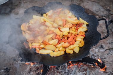Açık havada ateşte, dökme demir bir tavada pişen kızarmış patates ve havuçlar. Kamp ateşi hazırlama, duman, sıcak yağ ve geleneksel yemek malzemeleri, kamp, seyahat ve açık hava yaşam tarzı kavramları için uygundur..