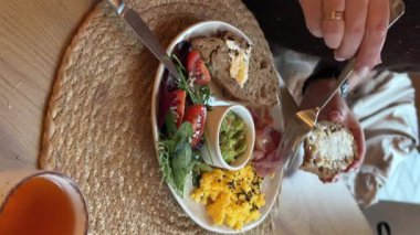 Besleyici bir kahvaltının dikey videosu. Bir insan omlet, guacamole ve sıcak bir kafe ortamında taze salata yanında tam tahıllı ekmek yemek için çatal kullanır..