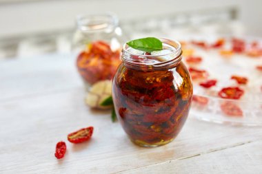 Ev yapımı güneşte kurutulmuş domatesler, beyaz ahşap zemin üzerinde cam bir kavanozda zeytinyağında sarımsak. Uzayı kopyala, kapat. Mutfak için yazdır.