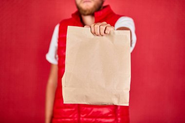 Kurye, kırmızı yelek giyen kurye kırmızı arka plandaki stüdyoda izole edilmiş kahverengi kağıt torbalarla online alışveriş yapıyor. 