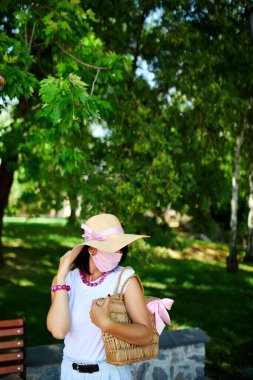 Hasır şapkalı ve çantalı bir kadın, pembe koruyucu maske takıyor, şehrin parkında, kişisel bakım kavramı, koronavirüs salgını sırasında hayat, covid-19.