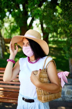 Hasır şapkalı ve çantalı bir kadın, pembe koruyucu maske takıyor, şehrin parkında, kişisel bakım kavramı, koronavirüs salgını sırasında hayat, covid-19.