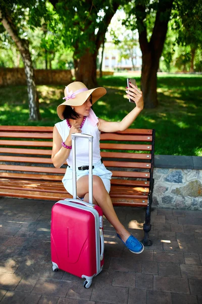 Blogger woman in straw hat and removed mask holds on her head, in the park outdoor with a suitcase, take selfie, life during coronavirus pandemic, opening air travel, travel concept.