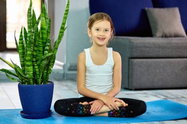 Mavi arka planda yoga sınıfında oturan bir kız. Evde eğitim, çocuk yogası, sakin ol, evde kal. Karantina, koronavirüs. Çocuklar evlerini karantinaya alırlar. Uluslararası yoga günü.