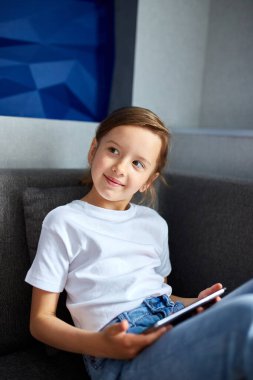Dijital tablet kullanan, kanepede oturan, oyun oynayan, gülümseyen ya da evde dizüstü bilgisayar başında çizgi film izleyen sevimli küçük bir kız. Modern çocuk ve eğitim teknolojisi.