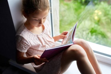 Evde, pencere kenarında kitap okuyan sevimli küçük bir kız evde ders çalışan bir çocuk, karantina, evde kalmak, yaşam tarzı....