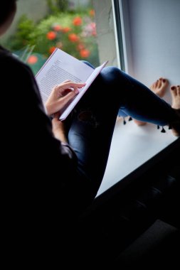 Evdeki pencerenin yanında kitap okuyan güzel bir genç kadın, pencere kenarında oturan bir kız, evde ders çalışan, karantina yapan, evde kalan, yaşam tarzı olan bir kız..