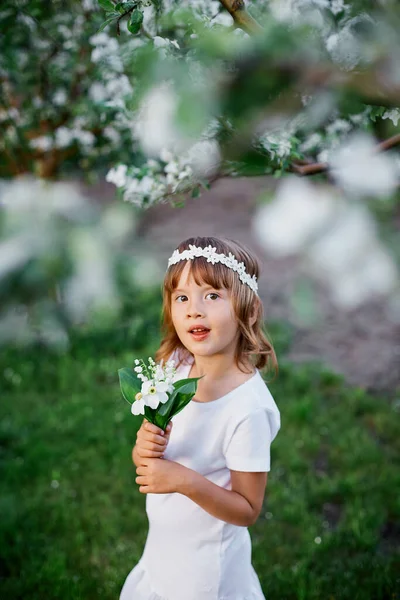 可愛い娘の肖像5 6歳の保有花春の庭に立って 白いドレスと花の花輪を屋外で身に着けて 春の季節が来ています — ストック写真