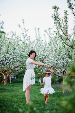 Mutlu kadın ve çocuk dansı, çiçek açan bahar bahçesinde güzel bir kız ve anne, açık havada beyaz elbiseler giyiyorlar, bahar sezonu yaklaşıyor. Anneler Günü Tatil Konsepti