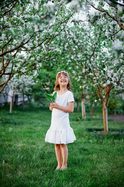 可愛い娘5 6歳の保有花春の庭に立って 白いドレスと花の花輪を屋外で身に着けて 春の季節が来ています — ストック写真