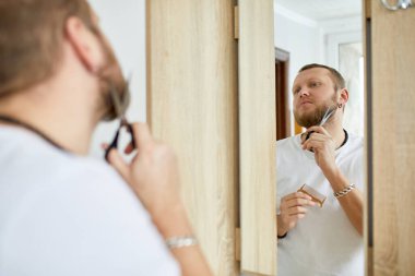 Beyaz tişörtlü yakışıklı adam sakal kesiyor, bıyığını evde makasla kesiyor. Karantina karantinası sırasında kişisel bakım. Evde kal..