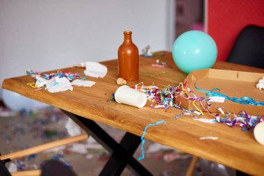 Parti kutlamalarından sonra parti kaosundan sonra parti artıklarını kutlamak için pizza kutusu, boş mavi şişeler, şarap bardağı ve parti süslemeleri olan dağınık ahşap masa.