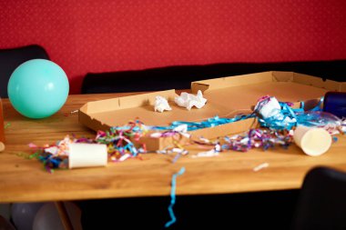 Parti kutlamalarından sonra parti kaosundan sonra parti artıklarını kutlamak için pizza kutusu, boş mavi şişeler, şarap bardağı ve parti süslemeleri olan dağınık ahşap masa..