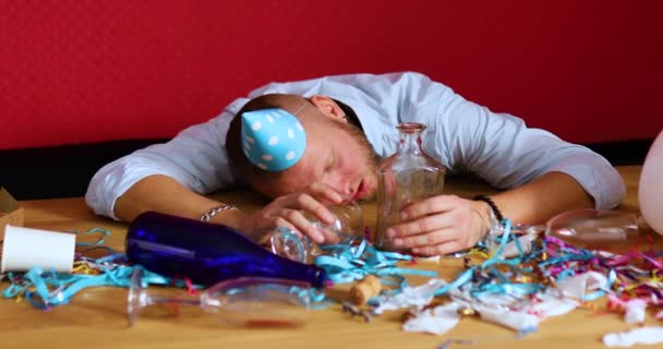Γενειοφόρος άντρας κοιμάται στο τραπέζι σε βρώμικο δωμάτιο με μπλε καπέλο μετά το μπάτσελορ ή το πάρτι γενεθλίων — Αρχείο Βίντεο
