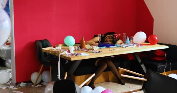 Mujer joven con lápiz labial rojo frotado y gorra, durmiendo en la mesa en la habitación desordenada después de la fiesta de cumpleaños — Vídeo de stock