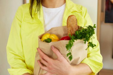 Sarı ceketli kadın, elinde taze meyve ve sebze dolu alışveriş poşetiyle evde duruyor, online alışveriş ve sağlıklı vejetaryen yeme kavramı dışında, mesaj için yer var.