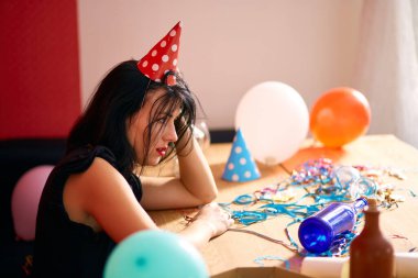 Kırmızı rujlu ve şapkalı genç bir kadın doğum günü partisinden sonra dağınık bir odada yorgun bir şekilde oturuyor.