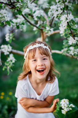 5-6 yaşlarında elinde çiçeklerle bahar bahçesinde dikilen beyaz elbisesi ve çiçekli çelengi olan komik bir kız çocuğu portresi geliyor..