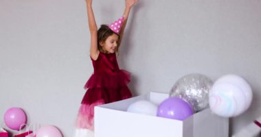 Pembe elbiseli ve şapkalı küçük neşeli kız doğum günü partisinde balonlarla hediye kutusunu açıyor, mutlu yıllar. Kutlama.