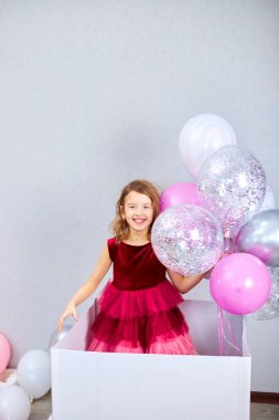 Pembe elbiseli şirin, neşeli küçük kız elinde balonlarla büyük hediye kutusundan dışarı bakıyor. Doğum günü partisi flamaları, doğum günün kutlu olsun. Kutlama.