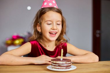 Küçük bir kız doğum günü şapkası takmış doğum günü pastasına bakıyor ve bir kutlama partisi konsepti için mum üflüyor..