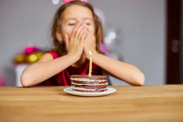 A little girl is wearing a birthday hat makes a wish , looking at a Birthday Cake, with glowing candles for a celebration party concept.