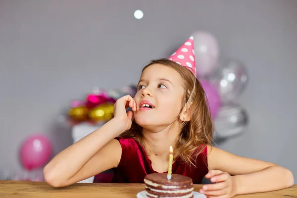 A little girl is wearing a birthday hat makes a wish , looking at a Birthday Cake, with glowing candles for a celebration party concept.