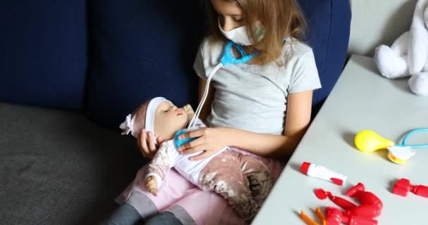 Mała dziewczynka w masce medycznej siedzi na kanapie, bawi się w lekarza, traktuje lalkę — Wideo stockowe