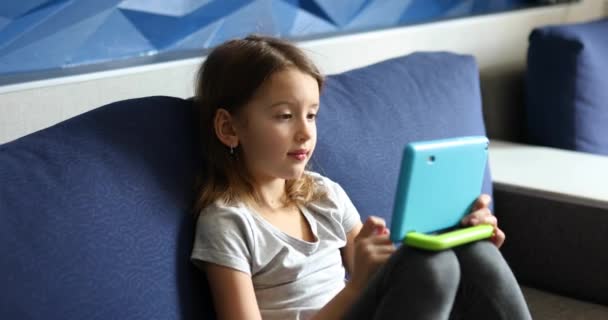 Симпатична дівчинка сидить на дивані, Кід залежний від технології, насолоджуючись грою онлайн на цифровому планшетному комп'ютері — стокове відео