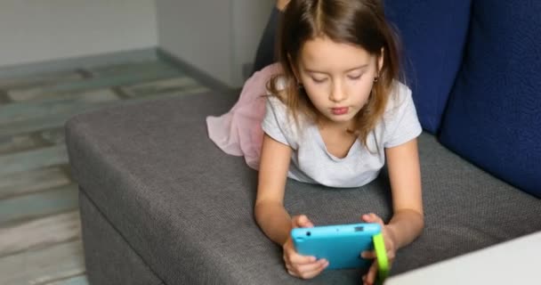 Мила маленька дівчинка лежить на дивані, Кід залежний від технології, насолоджуючись грою онлайн на цифровому планшетному комп'ютері — стокове відео