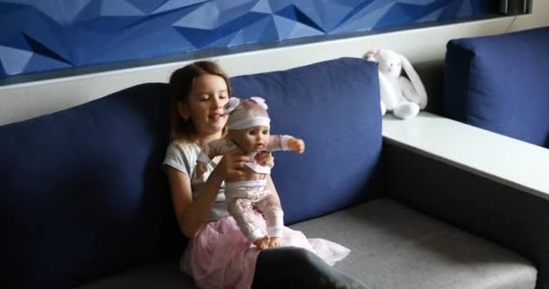 Liten flicka sitter på soffan i rummet hemma och leker med babydocka — Stockvideo