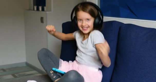 Щаслива маленька дівчинка в навушниках сидить і танцює на дивані, слухає музику, співає пісню — стокове відео