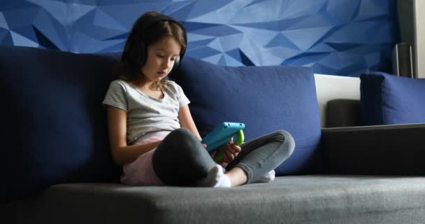 Cute little girl w słuchawki siedzi na kanapie z tabletem cyfrowym, grać w grę — Wideo stockowe