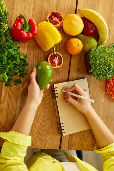 Kadın planlaması, yemek planlayıcısı üzerine haftalık yemekler yazması ya da evdeki mutfağında sağlıklı meyve ve sebzelerle ahşap masaya diyet planı yapması..