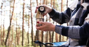 Genç bayan turist termosun açık olması ve sıcak içeceği çam ormanlarında bahar günü koltukta otururken bardağa doldurması, kadın gevşemesi ve sağlıklı yaşam tarzı konsepti, dışarı çıkın..