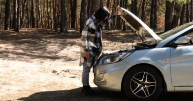 Genç bir kadın kaputu açıyor ve orman yolunda bozuk bir arabanın motorunu inceliyor.